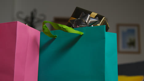 Close-Up-Of-Man-Putting-Down-Gift-Wrapped-Presents-In-Colourful-Bags-With-Tag-On-Table-In-Lounge-At-Home